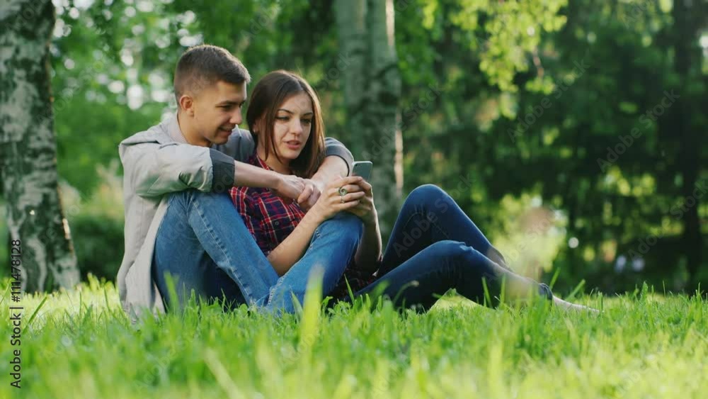 An attractive couple sitting on the grass in the park, use the phone, hugging An attractive couple sitting on the grass in the park, use the phone, hugging