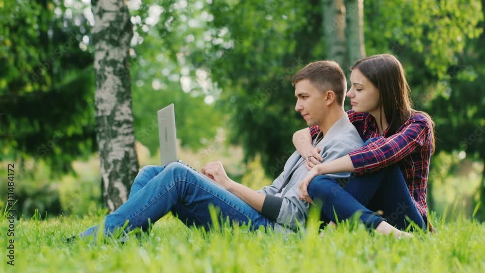 An attractive young couple sitting on the grass in the park, working with laptop. Girl embraces guy behind An attractive young couple sitting on the grass in the park, working with laptop. Girl embraces guy behind