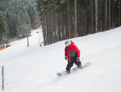 Male snowboarder slides down from the mountain in winter day, overlooking the snowy slope and forest at a winter resort