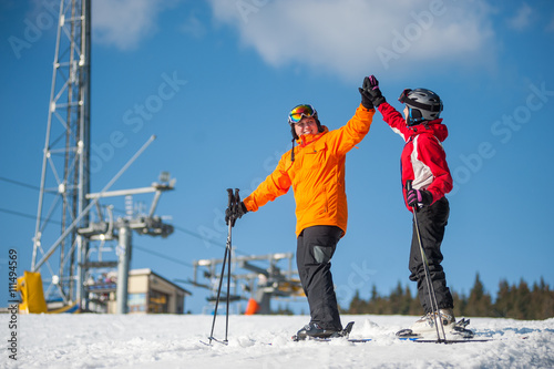 Man and woman holding hands, smiling looking at each other standing with skis on mountain top and giving each other a high five at a winter resort with ski lifts and blue sky in background