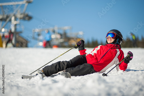 Woman wearing ski goggles, helmet, red jacket, gloves and pants lying with skis on snowy at mountain top and looking away in sunny day with ski lifts and blue sky in background.