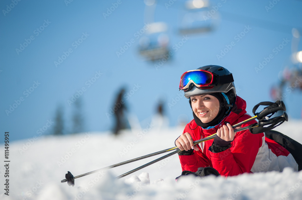Smiling woman wearing ski goggles lying with skis on snowy at mountain top and looking to the camera in sunny day with ski lifts and blue sky in background. Close-up
