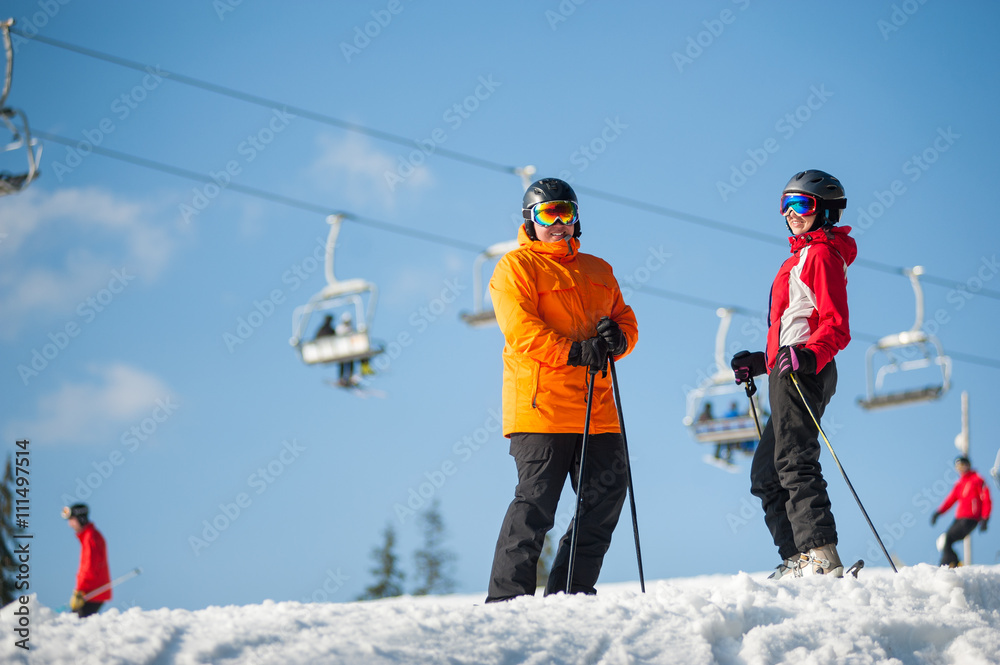 Happe couple man and woman wearing ski goggles standing with skis on mountain top at a winter resort in sunny day with ski lifts and blue sky in background.