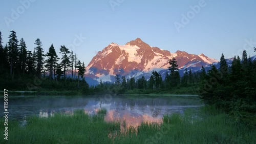 Dawn over the lake. Snowy mountain peak is reflected in the water. The forest on the lake. The singing of morning birds.