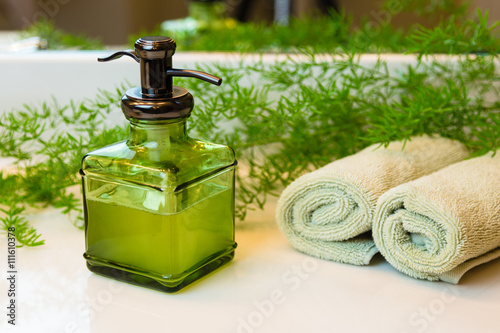 Pump green glass bottle with liquid castile soap. Rolled green towels in a spa setting. Green plant decor in background. Bathroom white countertop.