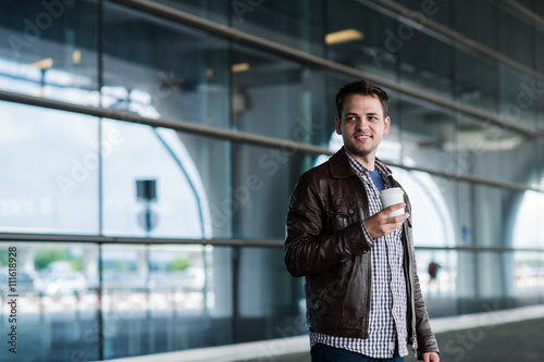 Stylish handsome young male traveller with bristle standing outdoors near the airport terminal. Man wearing jacket and shirt. Smiling person looking to camera holding cup of coffee