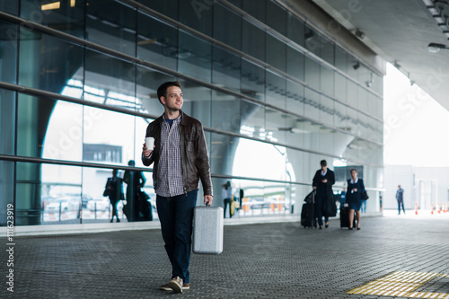Portrait of a young man walking near the airport with suitcase and coffee