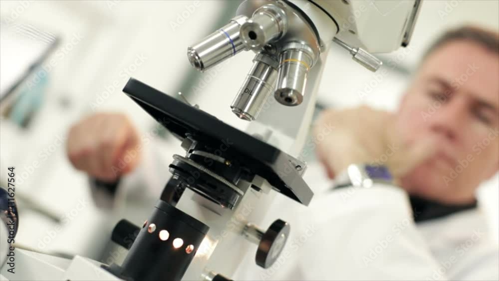A tilted frame shot of a modern microscope with a scientist who appears to be deep in thought out of focus in the background. Tilted framing