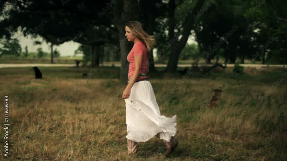 A lovely young woman wearing a long white skirt and cowgirl boots walks through a grove of trees on a windswept summer day then stop puts on her hat and looks around. A lovely young woman wearing a long white skirt and cowgirl boots walks through a grove of trees on a windswept summer day then stop puts on her hat and looks around.