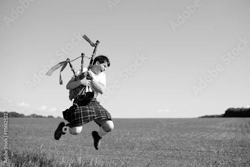 Black white photography of man jumping high with pipes in Scottish traditional kilt on summer field outdoors 
