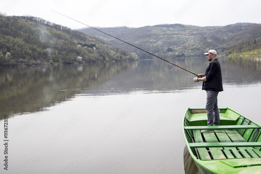 Naklejka premium Side View Of Sport Fisher With Cap And Vest Fishing From Green Canoe And Enjoying His Vacation On Beautiful Lake 