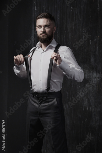 Attractive bearded man in a white shirt and suspenders standing near dark wall. He is strong, courageous and serious. He holds his suspenders. Dark room, night and harsh shadows.