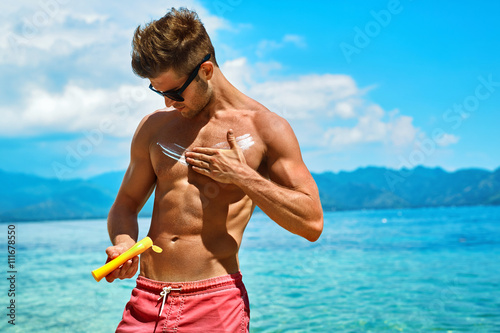 Man Skin Care In Summer. Handsome Male With Sexy Body In Sunglasses Applying UV Protective Sunscreen Lotion Before Sunbathing, Tanning At Beach Using Solar Sun Block Protection Cream For Healthy Tan.