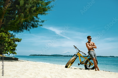 Handsome Muscular Man With Yellow Sand Bicycle Relaxing On Shore On Summer Travel Beach Vacation. Fitness Male Model With Bike Sunbathing By Ocean. Sports Activity And Recreation In Summertime Concept
