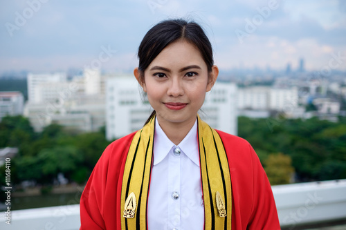 portrait of thai graduated young adult woman in red and golden uniform, polite and beautiful woman in uniform on graduation day, smiling standing in polite action