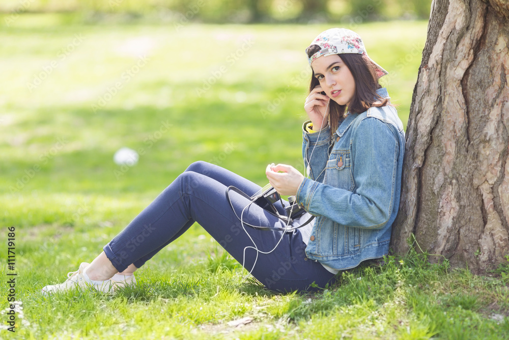 Teenage girl sitting in a park leaning on a tree and listening to music on headphones