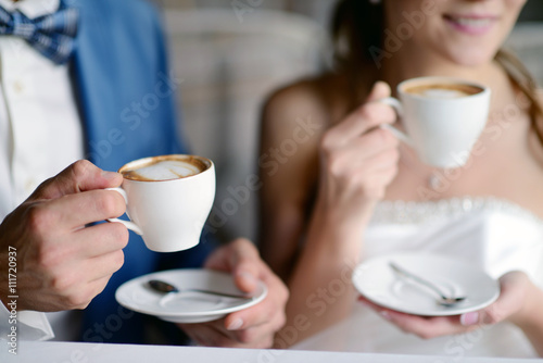 Wedding couple indoors is drinking coffee. Beautiful model girl in white dress. Man in suit. Beauty bride with groom. Female and male portrait. Woman with lace veil. Cute lady and handsome guy