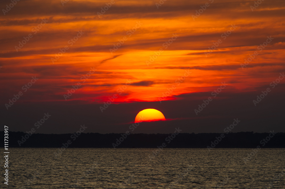 A large yellow sun slips into a bank of clouds as the sunset lights up a fiery orange sky.