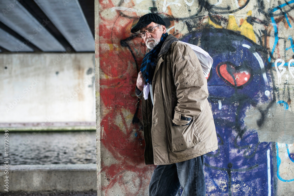 Homeless man leaning against concrete pillar under bridge.