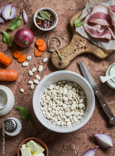 Ingredients for making bean mash with bacon - white beans, bacon, carrots, red onion, spices and herbs - . On a stone background. Top view