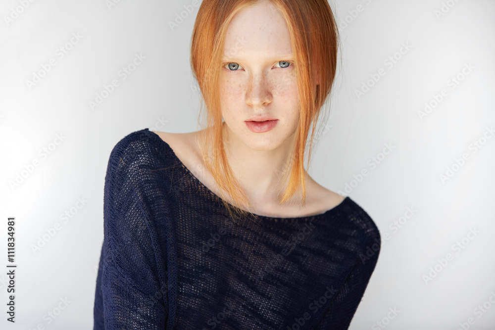 Beautiful serious Caucasian teenager with healthy freckled skin looking with thoughtful expression at the camera. Redhead girl wearing stylish black top posing isolated against white concrete wall