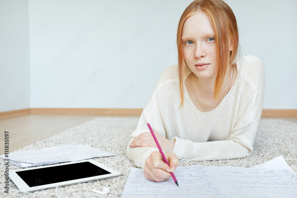 Attractive teenager with red hair and healthy freckled skin looking and smiling at the camera, while working on her course paper at home. Student girl using blank copy space tablet for doing homework Attractive teenager with red hair and healthy freckled skin looking and smiling at the camera, while working on her course paper at home. Student girl using blank copy space tablet for doing homework