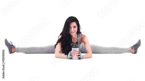 Beautiful sporty woman in grey overalls sitting in splits with shaker, Isolated over white background