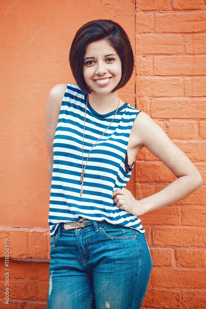 Portrait of beautiful smiling young latin hispanic girl woman with short hair bob, in blue jeans, striped tshirt, leaning on red brick wall in city looking in camera, toned with Instagram filters