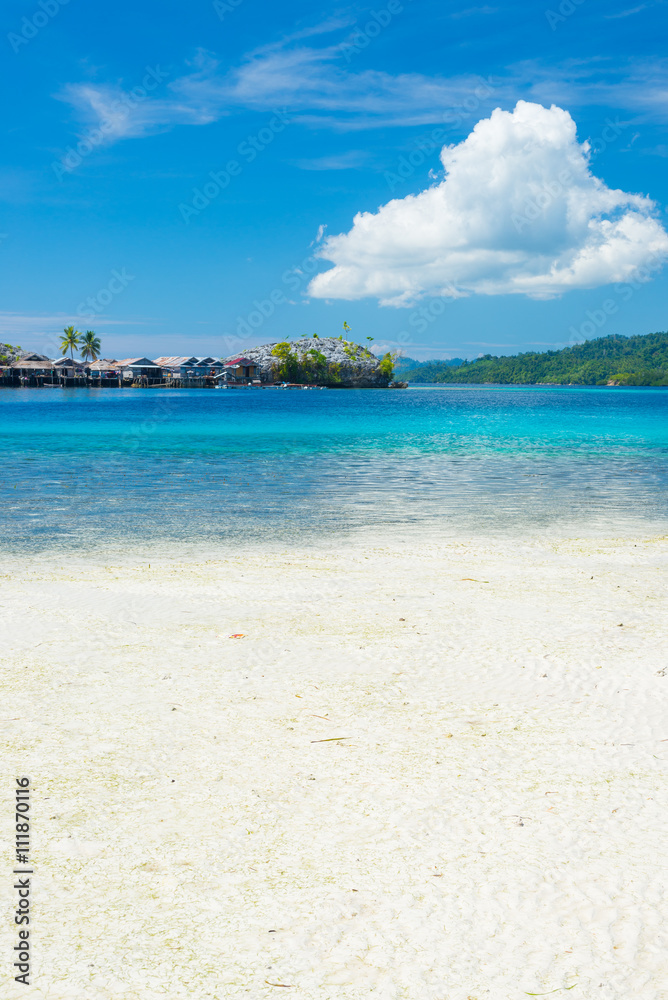 White sandy beach, turquoise transparent water and lush green jungle in the remote Togean (or Togian) Islands, Sulawesi, Indonesia. Islets and fishermen village in the background.