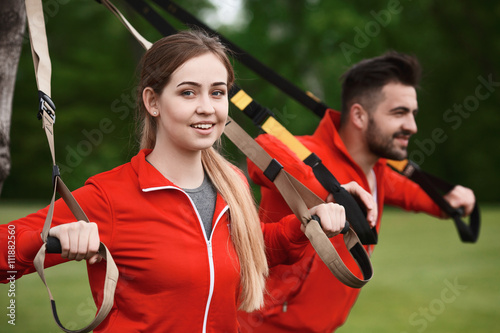 Closeup picture of beautiful sport or fitness woman training with suspension trainer sling with her colleague man in green park.