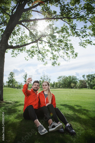 Sporty couple making selfies in green park or forest. Man and woman sittin on green grass and posing for photo camera.