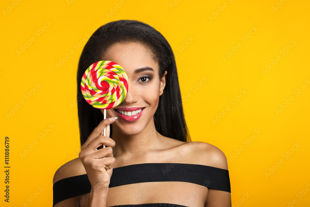 Smiling cute girl holding and covering her eye with big colourful sweet lollipop looking at camera over sunny, yellow background