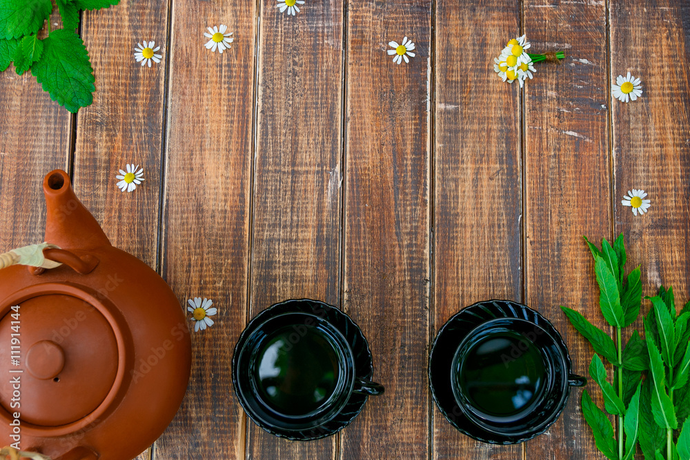 Brown teapot near two black cups of tea on wooden with fresh mint and chamomile table. Tea concept. Frame, copy space. Top view.
