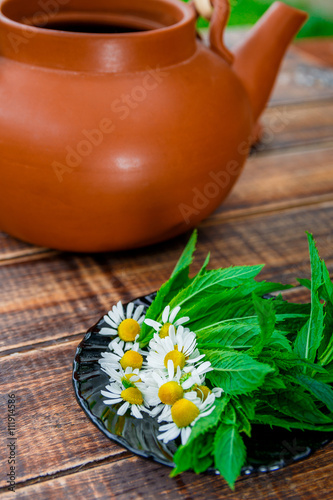 Open brown teapot with flower of chamomile near black plate with fresh mint and chamomile on wooden background. Closeup. Tea concept.
