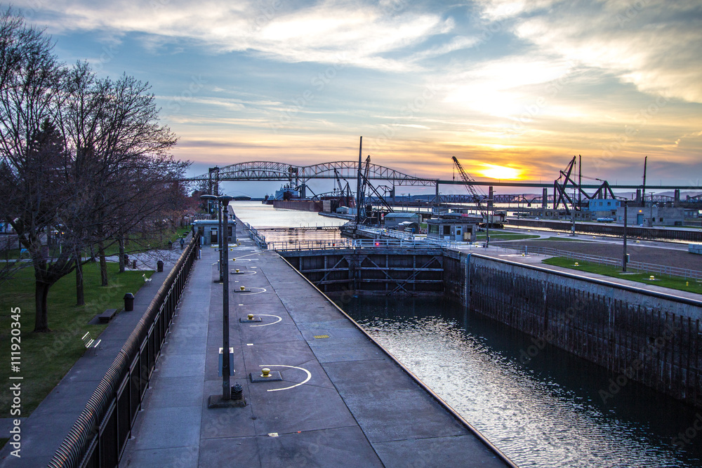 Naklejka premium Sunset Over The Sault Ste Marie Skyline And Soo Locks. The sunset over the Soo Locks and freighters with the International Bridge between the US and Canada at the horizon.