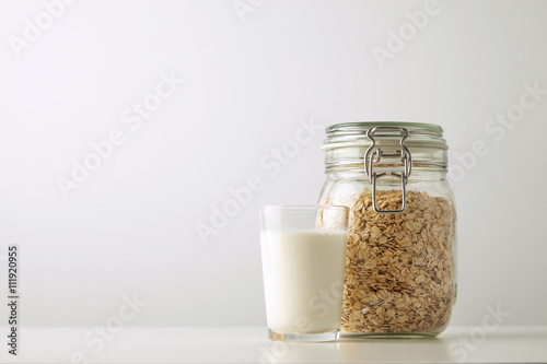 Transparent glass with fresh organic milk close to rustic jar with rolled oats isolated in side on white table