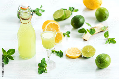 Home lime liquor in a glass and fresh lemons, limes on the white wooden background