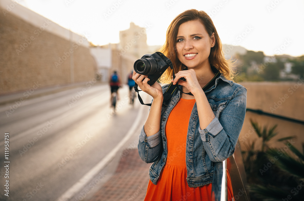 Outdoor summer lifestyle portrait of pretty, smiling, young woman having fun in the city, making photographs. Girl posing, looking into the camera lens. Concept travel photo of photographer shooting.
