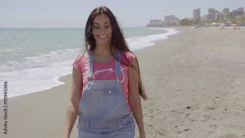 Single happy pretty woman in blue and pink top walking near beach with buildings in background Single happy pretty woman in blue and pink top walking near beach with buildings in background