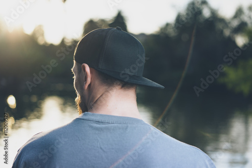 Photo Bearded Young Man Wearing Black Blank Cap. Green City Park Lake Background at Sunset. Relaxing time after hard work day. Back view. Horizontal Mockup.