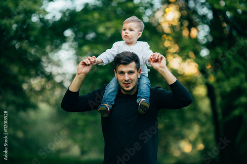 happy little boy stretching out hands while his father carrying him on shoulders in park