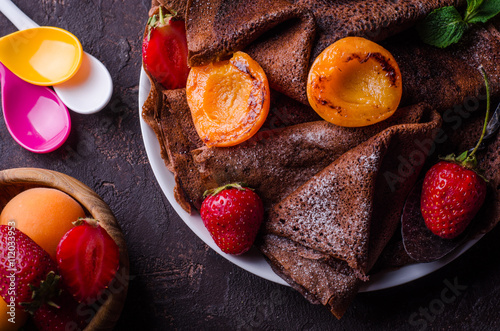 Stack of chocolate crepes decorated with strawberry and grilled apricots on dark slate background. Selective focus. Top view. Background with place for some text