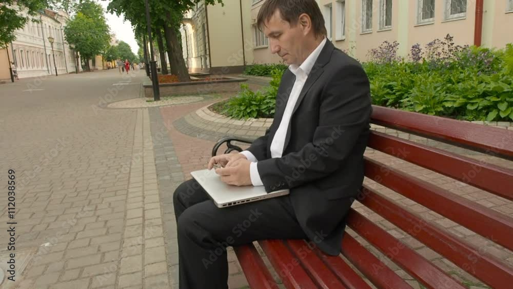 Businessman tweeting on cellphone sitting on a bench in the street Businessman tweeting on cellphone sitting on a bench in the street