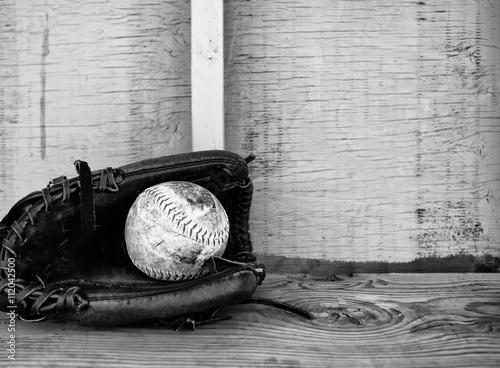 A softball laying in the pocket of a ball glove on a wood bench in black and white photo