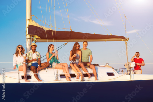 Smiling people on small yacht. Guys and girls holding drinks. Captain is watching the horizon. Perfect weather for sailing. © DenisProduction.com