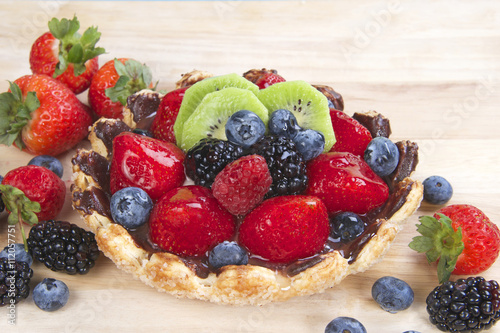 Fresh fruit tart with flower shaped pie crust sprinkled with sugar crystals coated with chocolate filled with custard topped with fresh strawberries, blue berries, boysenberries and kiwis