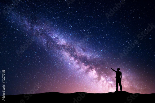 Milky Way with silhouette of a standing young man pointing finger in night starry sky on the mountain. Night landscape. Beautiful Universe, travel background with purple sky full of stars and light