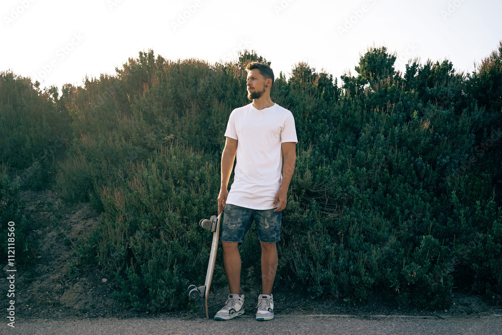 Fototapeta premium Young male wearing a blank white t-shirt and shorts is looking aside. A skater with a board is standing on a bush wall background.