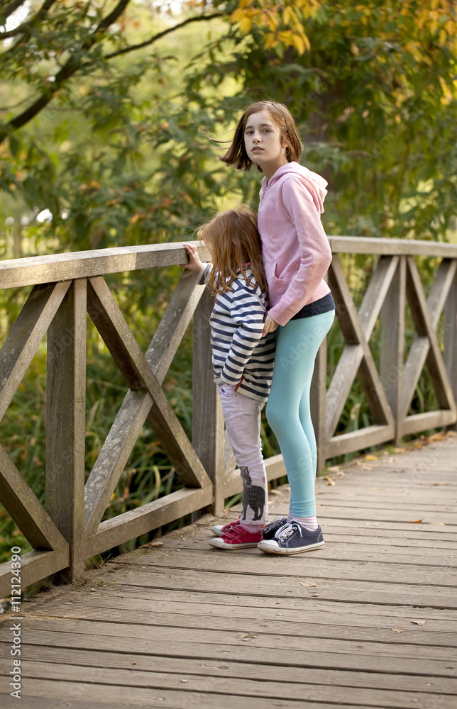 Sisters. A young girl keeps an eye out for her younger sister as they stand together on a bridge in a local park watching the ducks.