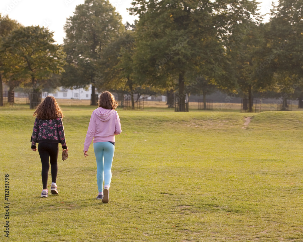 Fototapeta premium Heading Home. Two young sisters walk together towards the exit of the park. The sun is setting and the low afternoon light is outlining their figures.